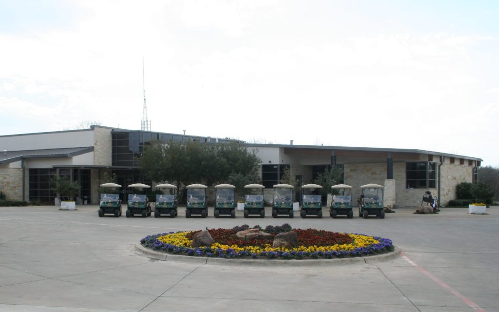A row of golf carts parked in front of the Bridges Clubhouse at Firewheel Golf Park, featuring a distinctive circle of greenery that resembles the Firewheel logo